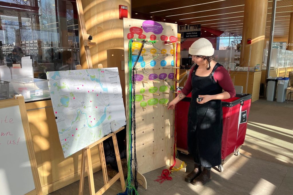 A person stands next to a board with colorful string that helps collect demographic data from the user. Next to the board is a map with dots on it.