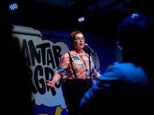 Logan Lopez, a poet with large glasses and a backwards orange ball cap stands at a microphone in low, blue light wearing a pink shirt with multi-colored dinosaurs on it.