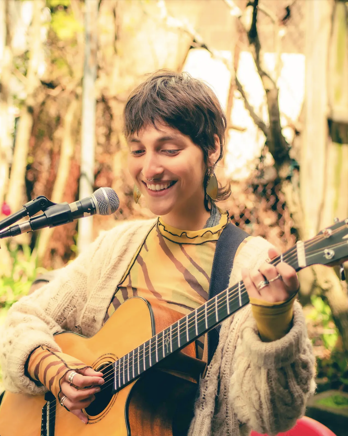 Mel, a white nonbinary person with a shaggy mullet smiles and plays guitar while singing into a microphone on a sunny day with trees in the background