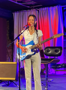 Mada Silva, a black woman with long curly hair dress in a monochromatic beige outfit holding a royal blue electric guitsr