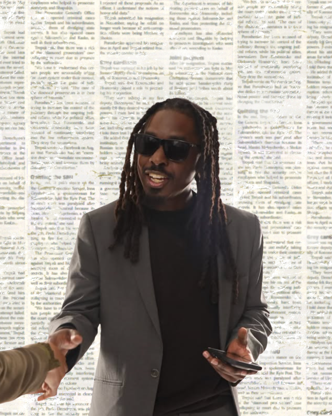 John Kind, a black American man with dreadlocks past his shoulders wearing a gray suit jacket and black t-shirt with sunglasses on a newsprint background