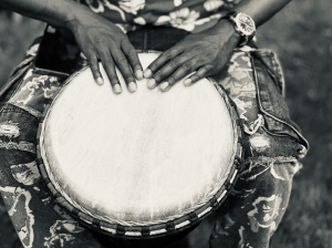 a bongo drum suspended between a person's knees with their hand touching the top