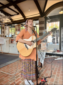 Mel Starr, a white, nonbinary singer/songwriter wearing an orange sweater and long patterened skirt plays the guitar and sings into a microphone