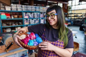 Alex Adamo, a woman with short black bangs, long hair with green dip dyed edges, holding a basket filled with multicolored felt yarn