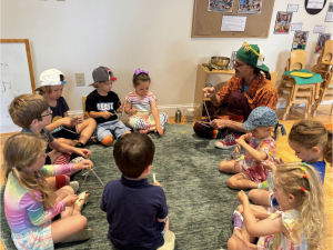 Rafael Medina, wearing a bright green backwards baseball cap with yellow sunglasses perched on the rim, sits on the floor surrounded by children playing the triangle
