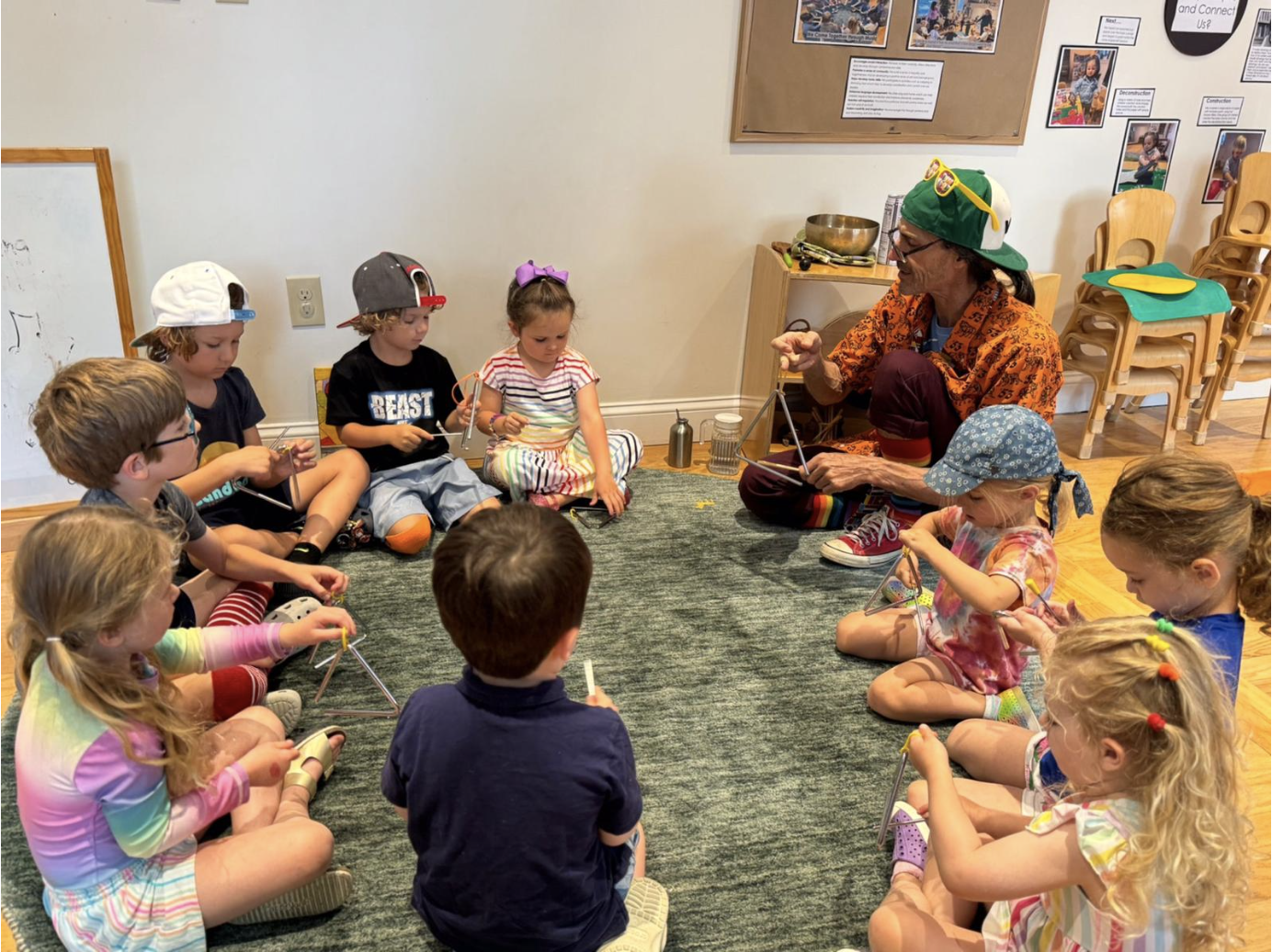Rafael Medina, wearing a bright green backwards baseball cap with yellow sunglasses perched on the rim, sits on the floor surrounded by children playing the triangle