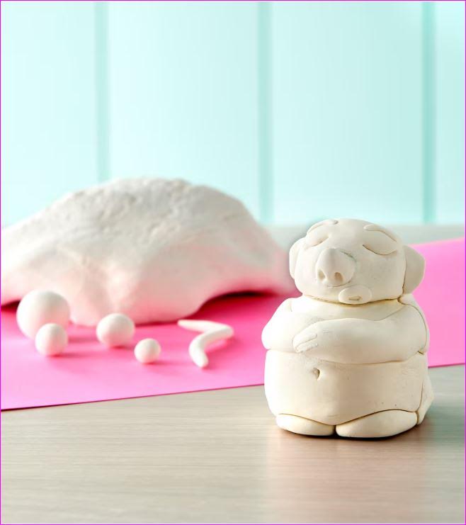 a table scape featuring a big lump of air dry clay and pieces of clay rolled into balls atop a pink tablecloth. There is a small person with their arms crossed sculpted out of clay in the foreground.