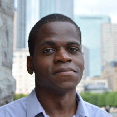 Merlo looking at the camera wearing a blue button up shirt in front of a city backdrop