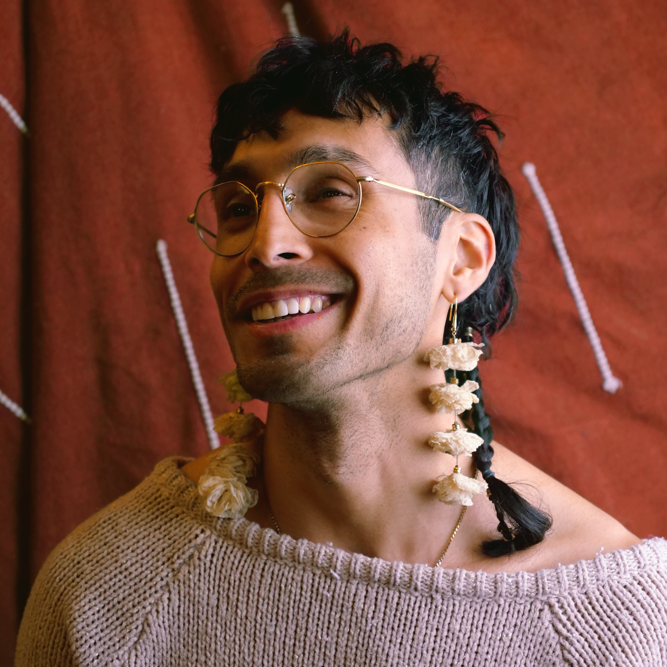 Paxtī smiling with floral earrings and a pink sweater in front of a red backdrop
