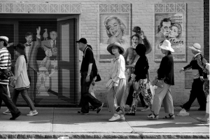 a group of tourists, most wearing sun hats, walk by the famous Harvard Sq. theater walls with murals of marilyn monroe and charlie chaplin in the background
