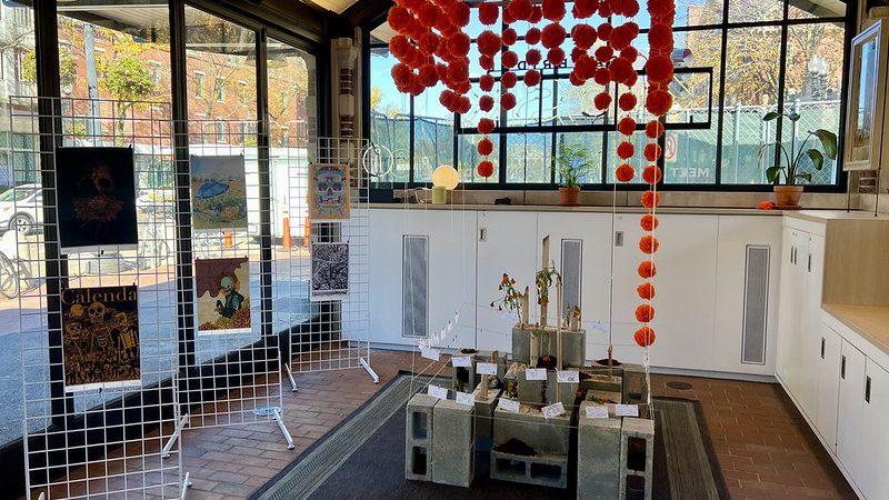 A hanging mobile of bright orange marigold flowers hands over a cinderblock structure. Art prints hang on wire grids on the left. 