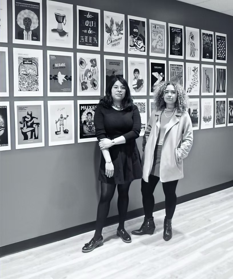A black and white photo of Sitalin Sánchez (left) and Michelle Segura (right) standing in front of a poster-covered wall.