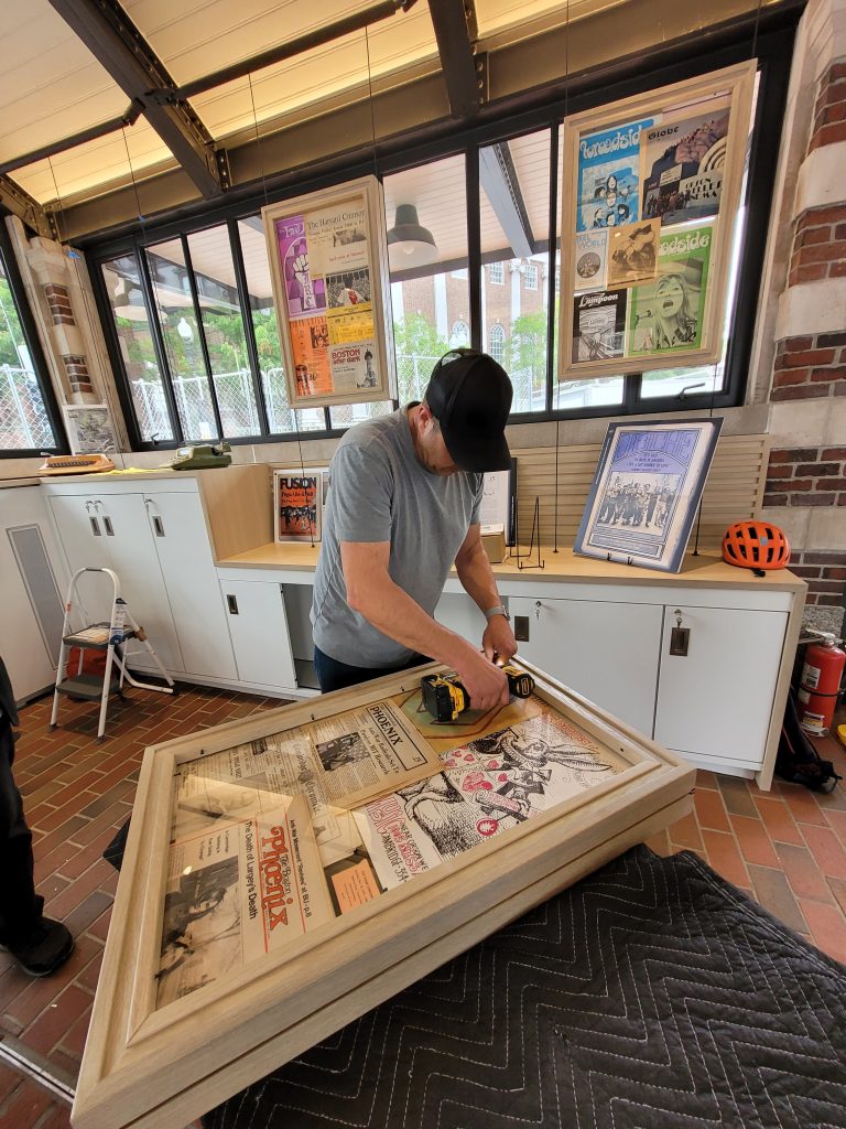 Someone drills shut a thick wooden frame displaying iconic newspaper front pages while setting up for the Cambridge: Then and Now exhibition.