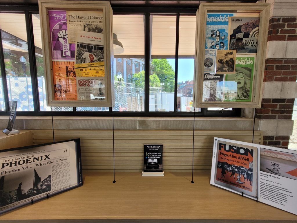 Colorful front pages from old newspapers hang in floating frames as part of the Cambridge: Then and Now exhibition. Other framed images sit on the shelf below.