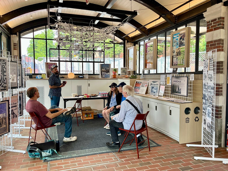 Visitors at the KiOSK sit on red chairs and talk, surrounded by white wire grids with posters. There is a table with snacks and refreshments at the back.