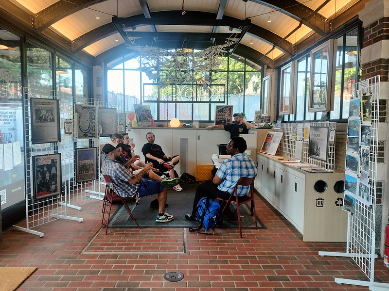 Visitors at the KiOSK sit on red chairs and talk, surrounded by white wire grids with posters.