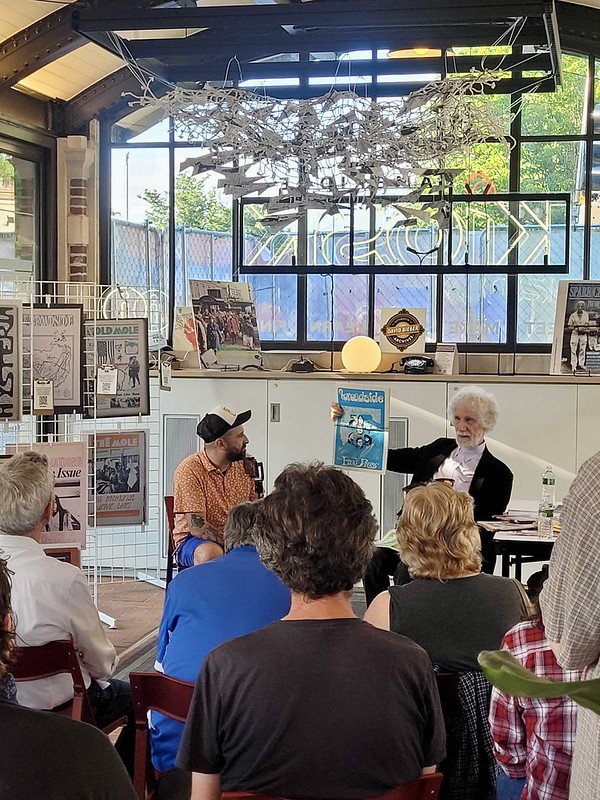 Two people speak in front of an audience with their backs to the camera. One holds up a piece of newsprint as he talks.