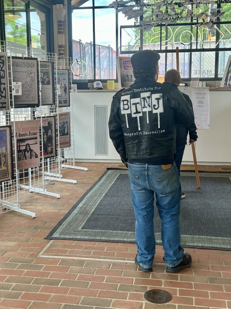 A visitor wearing a leather jacket with the Boston Institute for Nonprofit Journalism's logo on it stands with their back to the camera, looking at the exhibition created by that same organization.
