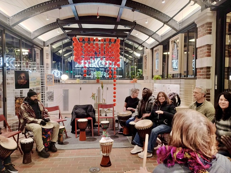People smile and play in a community drum circle under hanging chains of paper marigolds