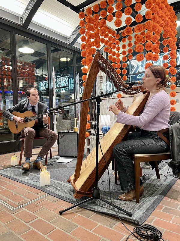 One performer plays guitar on the left, while another sings and plays the harp on the right. Images of Mexico's Festival Mictlán exhibition is visible in the background.