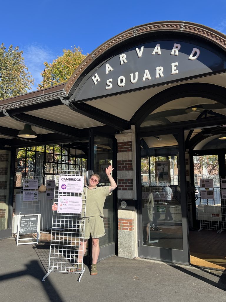 Someone holds a wire frame and smiles in front of the KiOSK during installation of the Queer History exhibition.