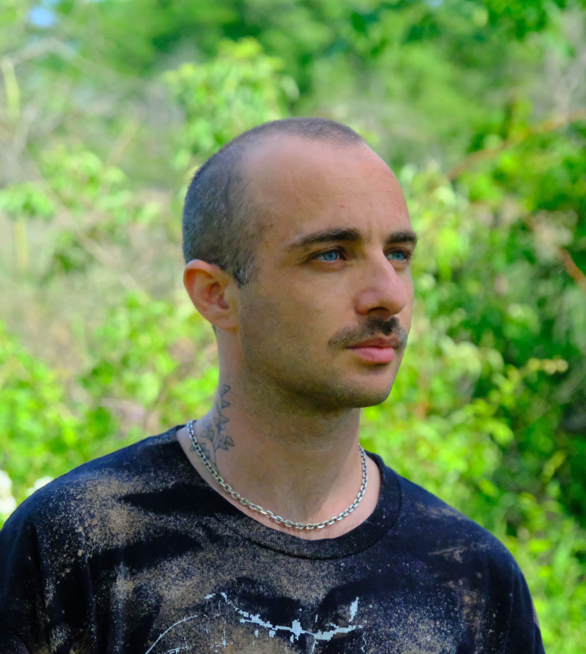Image shows a blue-eye white-presenting adult with a mustache and shaved head starring off into the distance. In the background are green leaves.