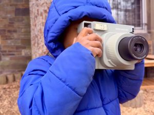 Picture features a child in a hooded blue winter coat holding a large white portable camera with both hands.