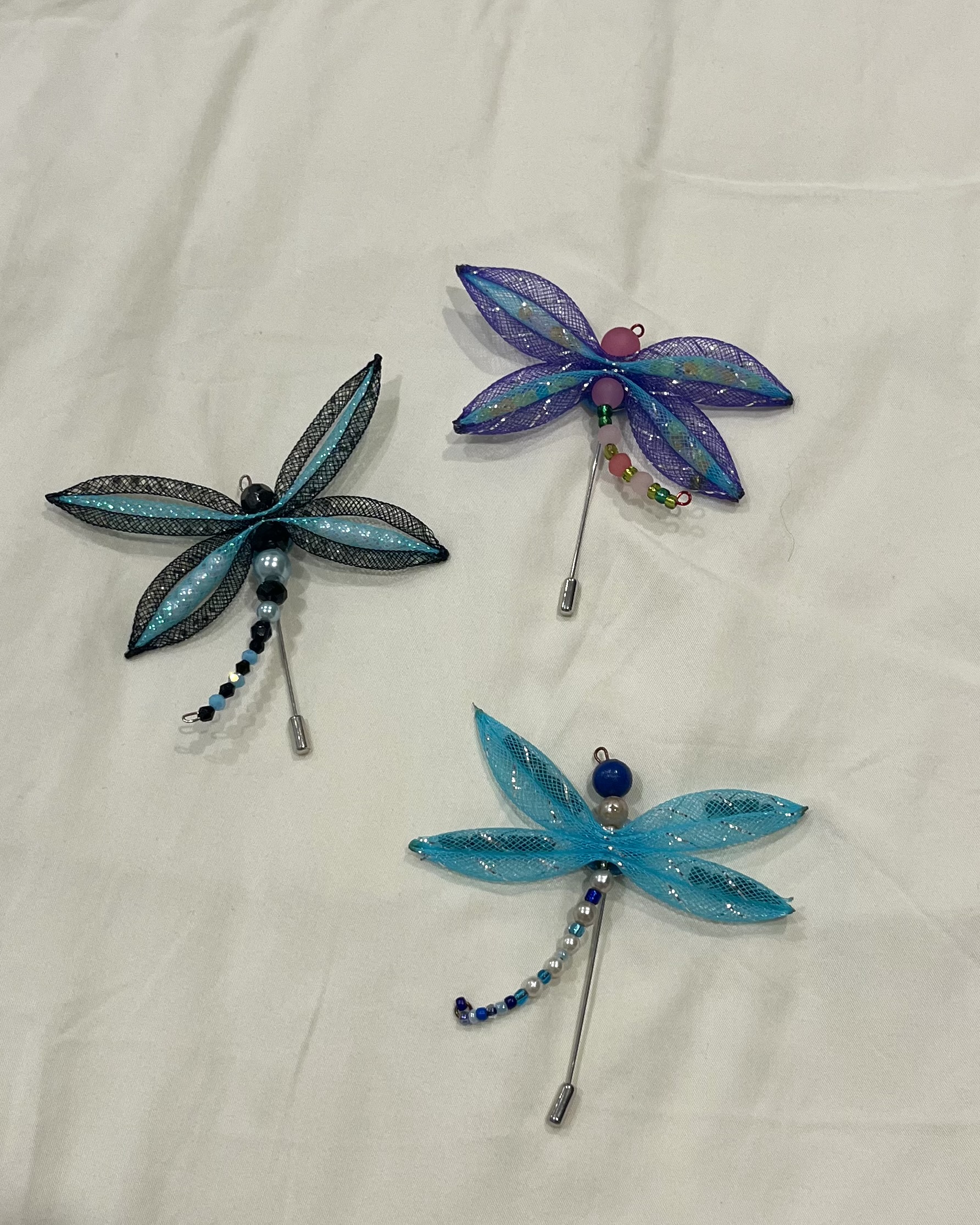 Three dragonflies made of beads and mesh placed on an off-white cloth